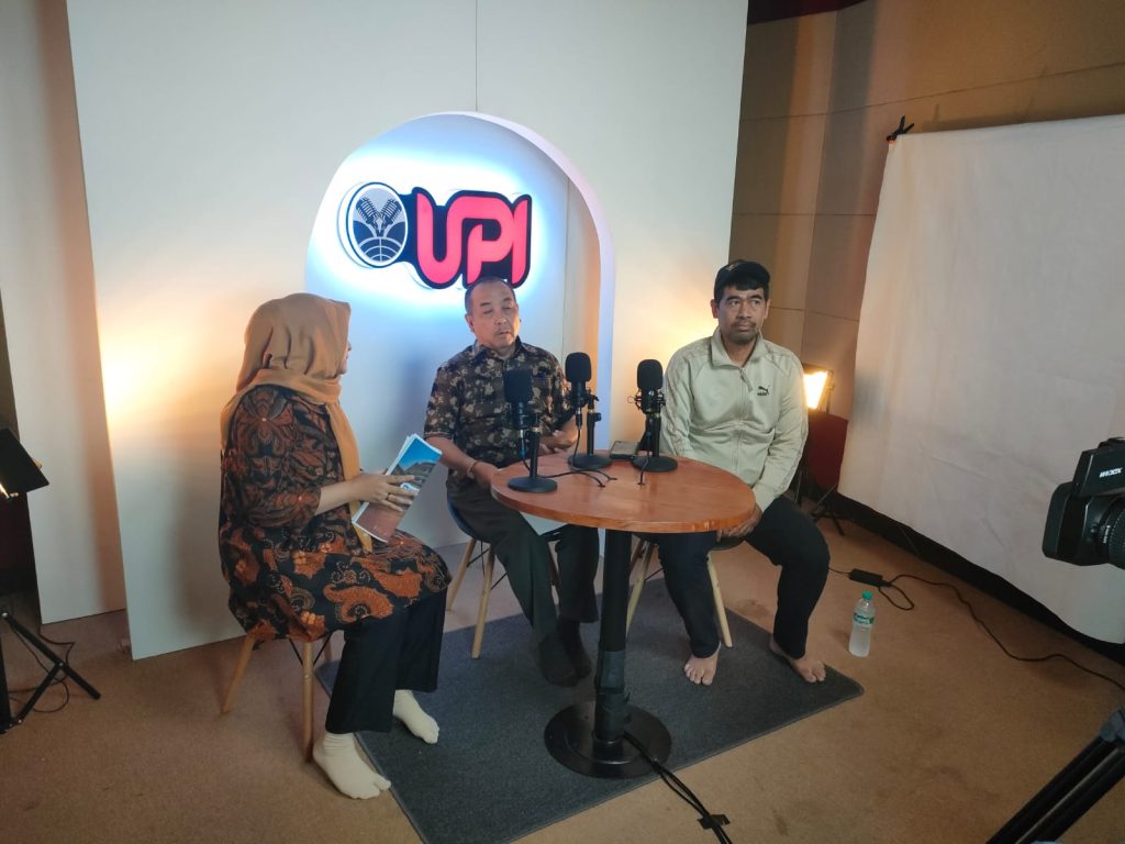 Tiga orang sedang duduk di depan meja kecil dengan mikrofon, siap untuk melakukan rekaman atau wawancara. Orang yang berada di tengah adalah Rektor Universitas Pendidikan Indonesia (UPI). Di belakang mereka, terdapat logo UPI yang menyala, dan suasana studio terlihat dengan pencahayaan serta peralatan kamera.